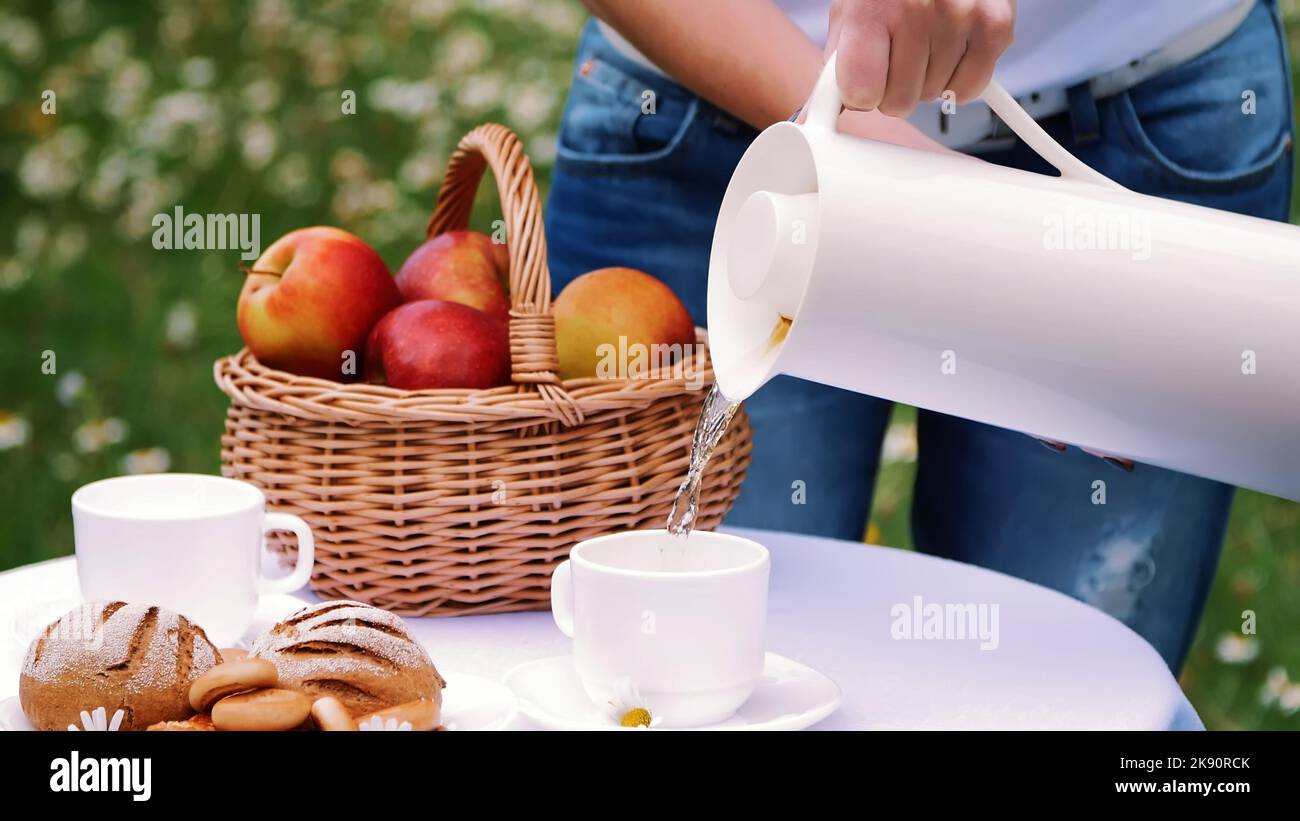 On the background of a chamomile lawn, women's hands pour tea from a white jug into a white cup, a thermos bottle. On the table there is also a basket with red apples. High quality photo Stock Photo