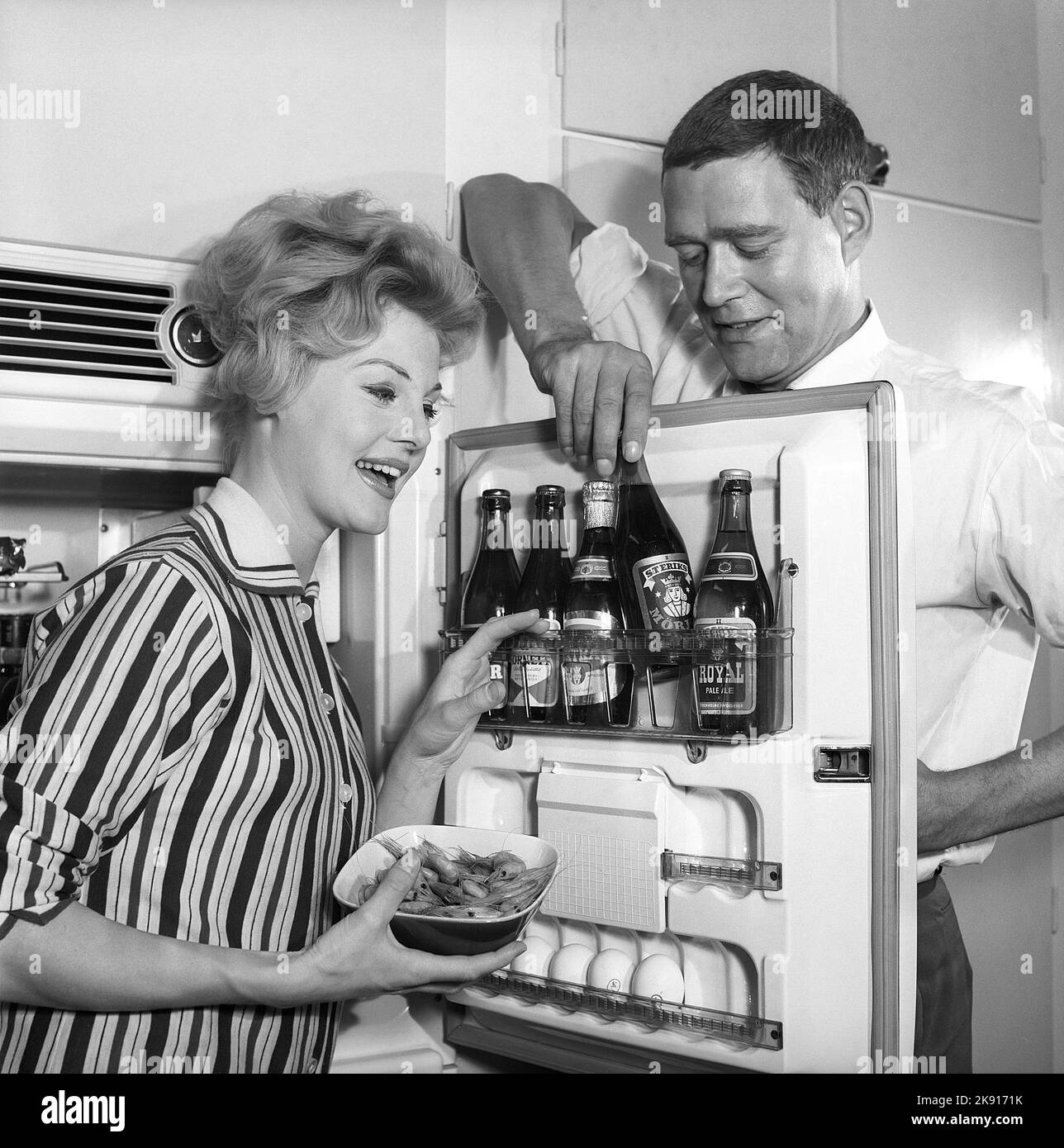 In the kitchen 1950s. A couple in their kitchen and at the refrigerator where the beer is kept in the door shelf. Five beer bottles are seen. Mrs is holding a bowl of shrimps. Sweden 1959. Kristoffersson ref CH77-1 Stock Photo