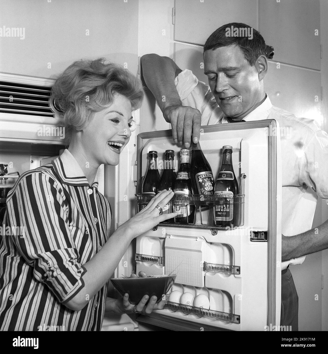 In the kitchen 1950s. A couple in their kitchen and at the refrigerator where the beer is kept in the door shelf. Five beer bottles are seen. Mrs is holding a bowl of shrimps. Sweden 1959. Kristoffersson ref CH77-3 Stock Photo
