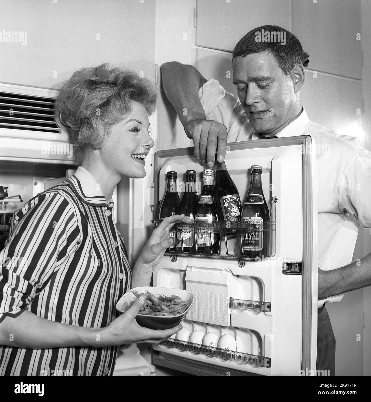 In the kitchen 1950s. A couple in their kitchen and at the refrigerator where the beer is kept in the door shelf. Five beer bottles are seen. Mrs is holding a bowl of shrimps. Sweden 1959. Kristoffersson ref CH77-2 Stock Photo