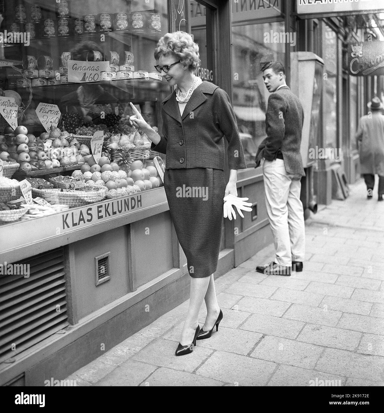 In the 1950s. A woman is pictured in the street and at the storefront of a fruit and groceries store. She looks as if she is deciding what to buy. Dressed in a matching jacket and skirt.  Sweden 1959 Kristoffersson ref CF58-4 Stock Photo