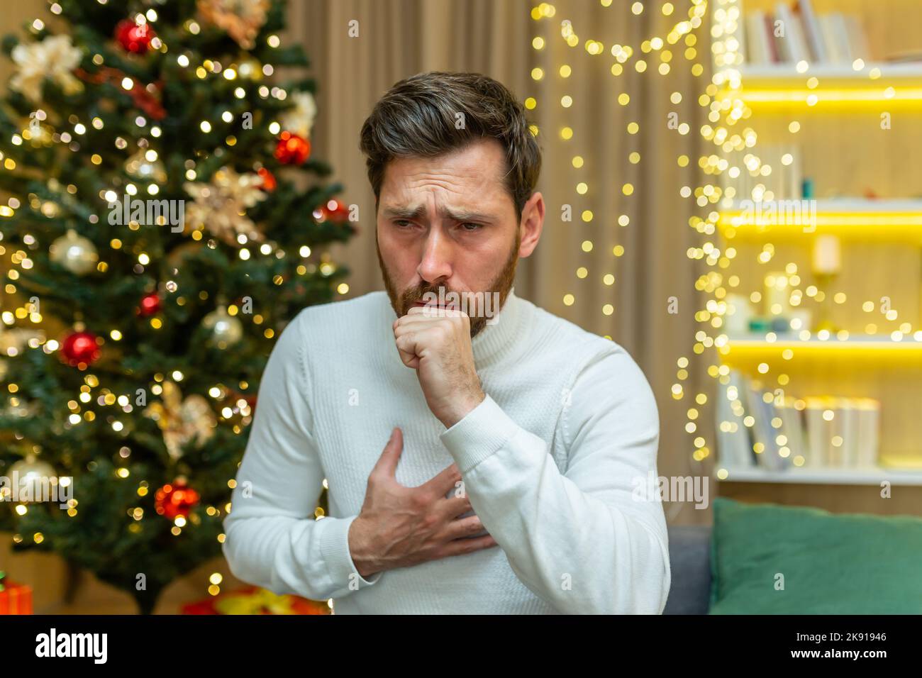 man coughs on christmas at home sitting alone on sofa sick on new year holidays in living room. Stock Photo