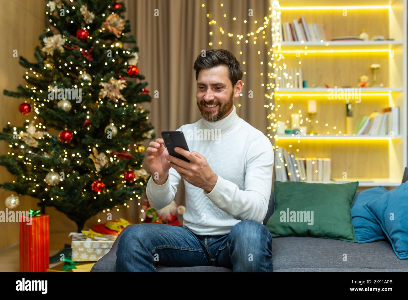 Cheerful man at home uses the phone, celebrates Christmas and New Year sitting on the sofa near the Christmas tree, types messages and browses online on the Internet. Stock Photo