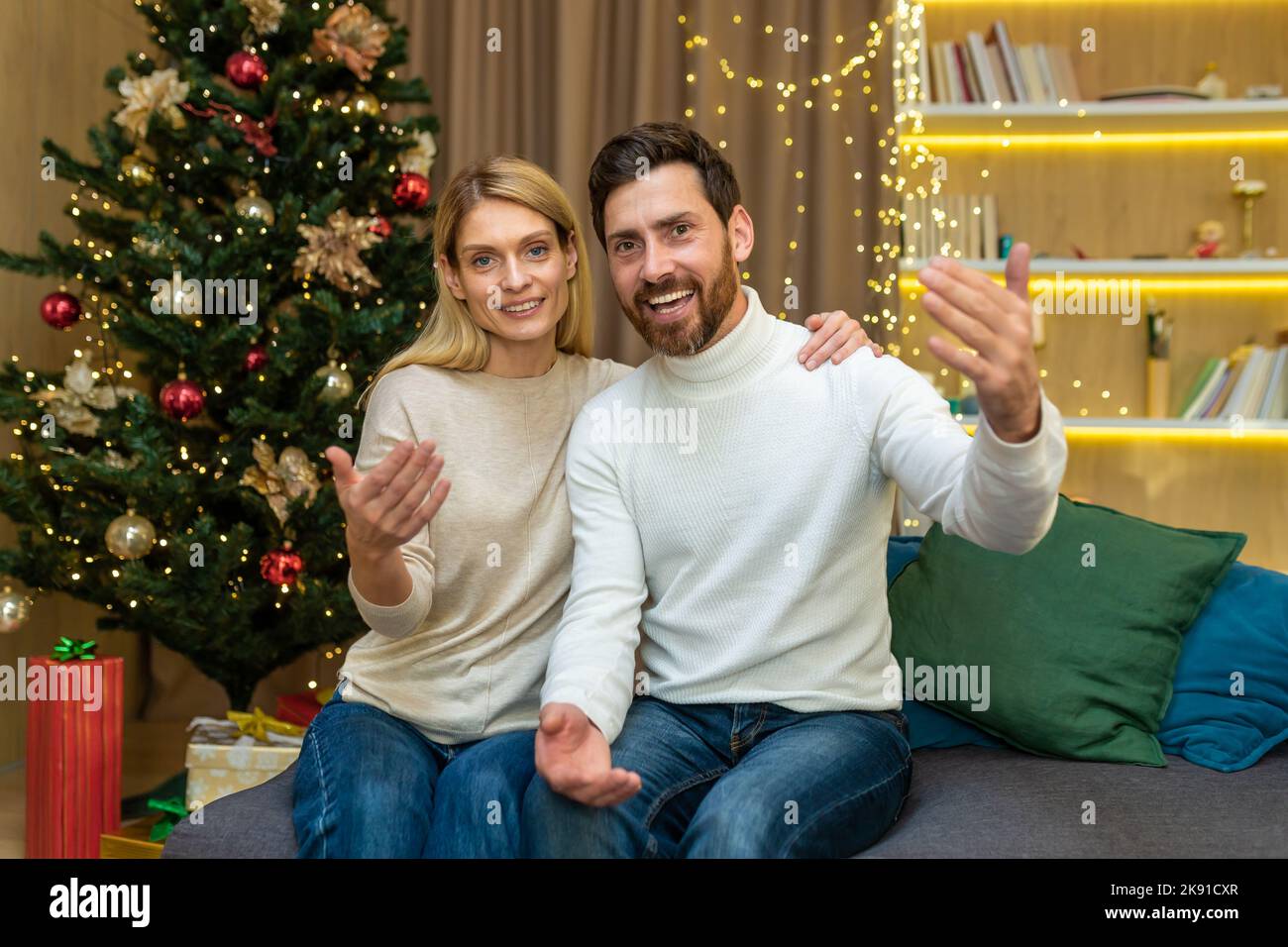Video call family couple, a man and a woman, are talking to friends, remotely looking at the web camera, and waving and greeting, inviting friends to visit to celebrate Christmas and the New Year. Stock Photo