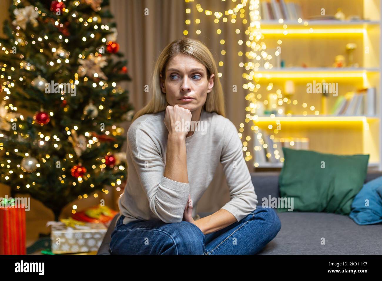 sad Woman alone at home for Christmas and New Year, sitting depressed near Christmas tree on sofa in living room, waiting for celebration. Stock Photo