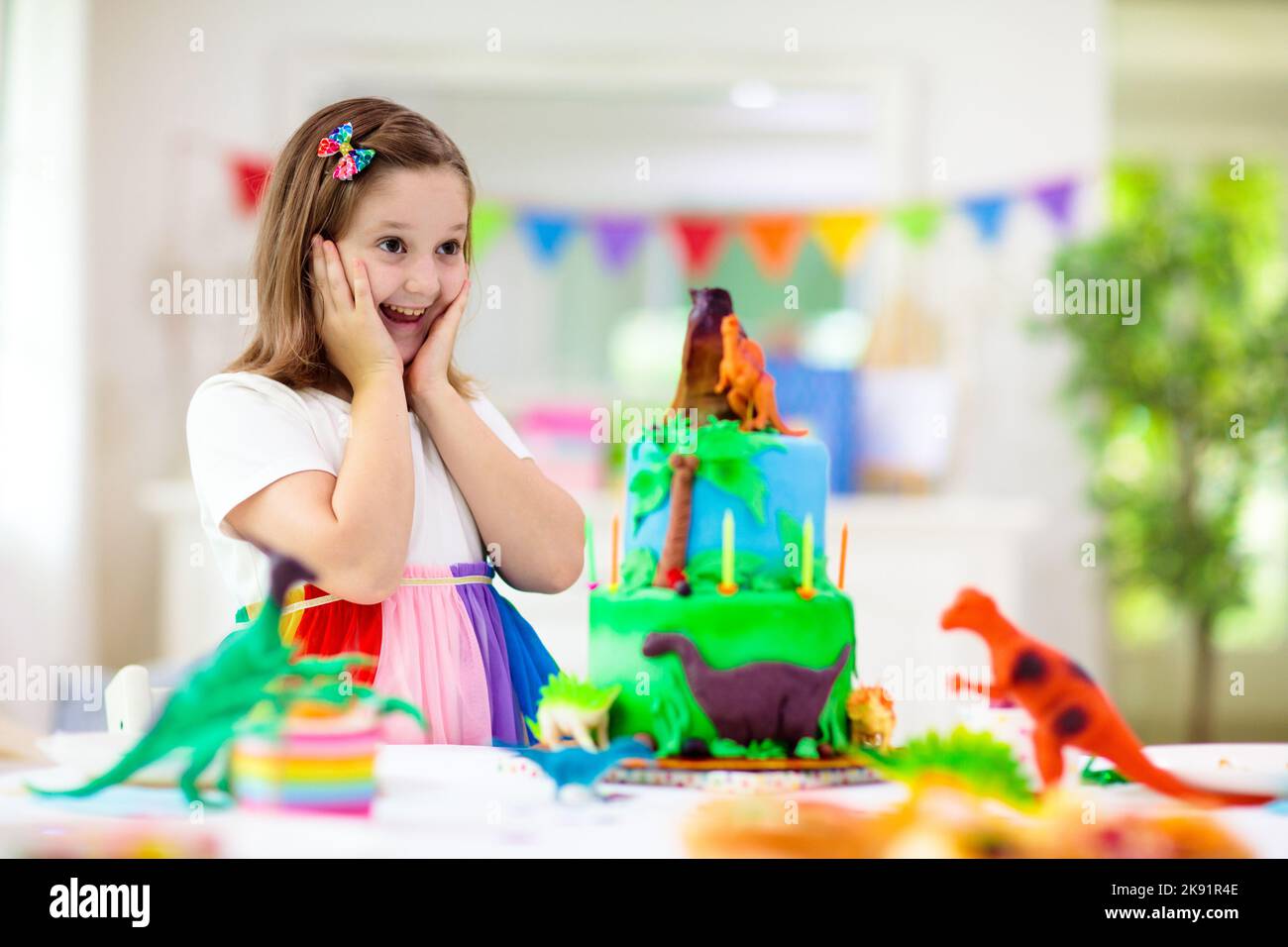 Kids birthday party. Dinosaur theme cake. Little girl blowing candles and opening gifts. Children event. Decoration for dinosaurs themed celebration. Stock Photo