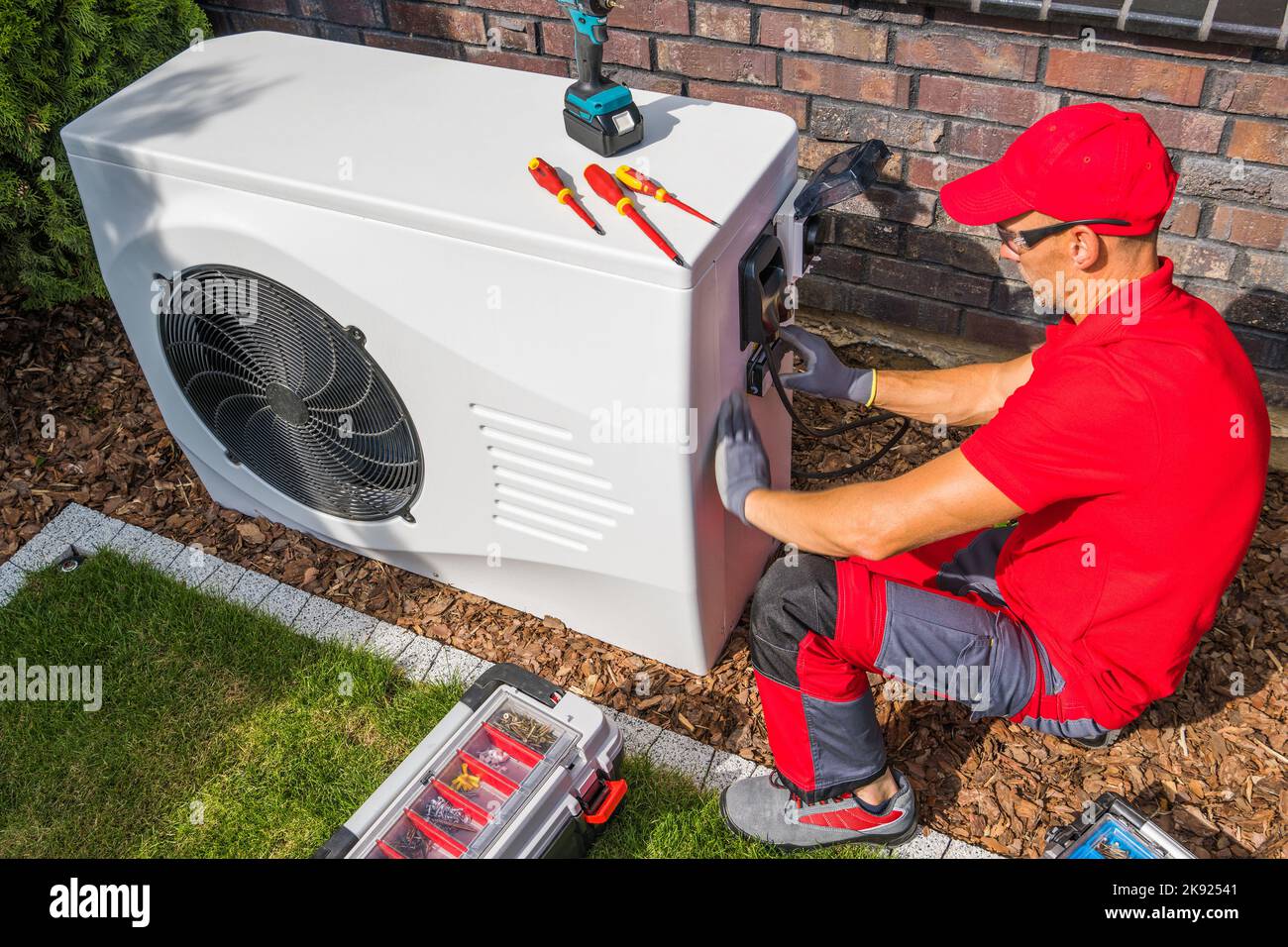 Professional Middle Aged HVAC Technician in Red Uniform Repairing Modern Heat Pump Unit. House