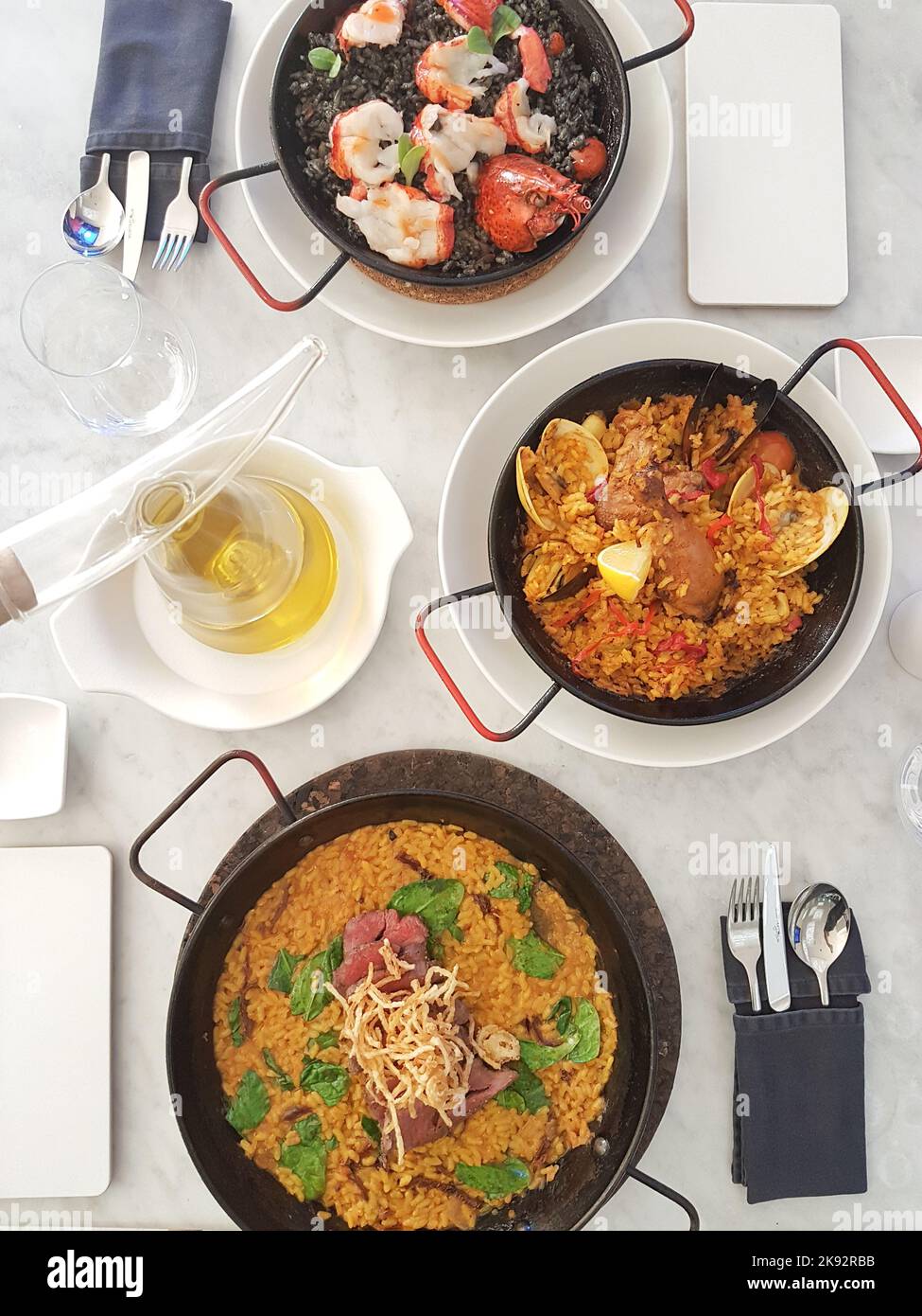 Flatlay of three solo Spanish paella dishes. Paella Negra, Valencia, Chicken on white marble table. Cutlery in table napkin, olive oil in glass bottle. Stock Photo