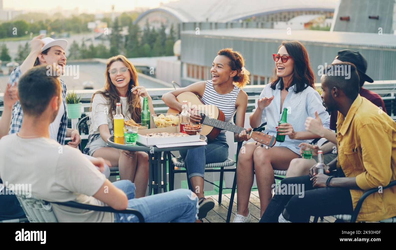 Pretty African American girl is playing the guitar while her happy friends multiethnic group are singing and moving hands enjoying party on rooftop. Table with food and drinks is visible. Stock Photo