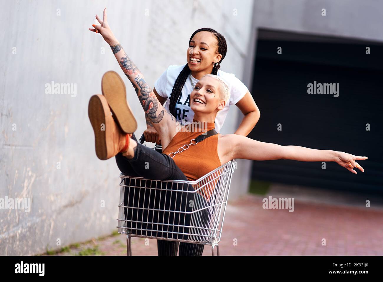 Im having good fun. an energetic young woman pushing her female friend in a shopping cart outdoors. Stock Photo