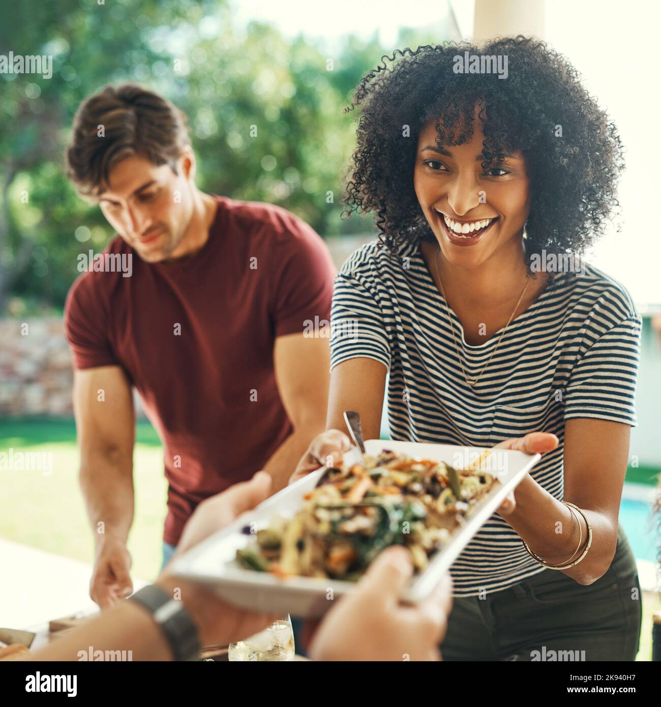 Good food always brings everyone together. a beautiful young woman passing over a plate of food at a family gathering outdoors. Stock Photo