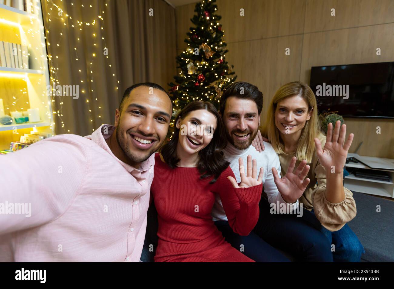 Diverse friends celebrating christmas and new year together, group of people having dinner at home sitting on sofa near Christmas tree, taking selfie photo and talking with friends on video call using phone. Stock Photo