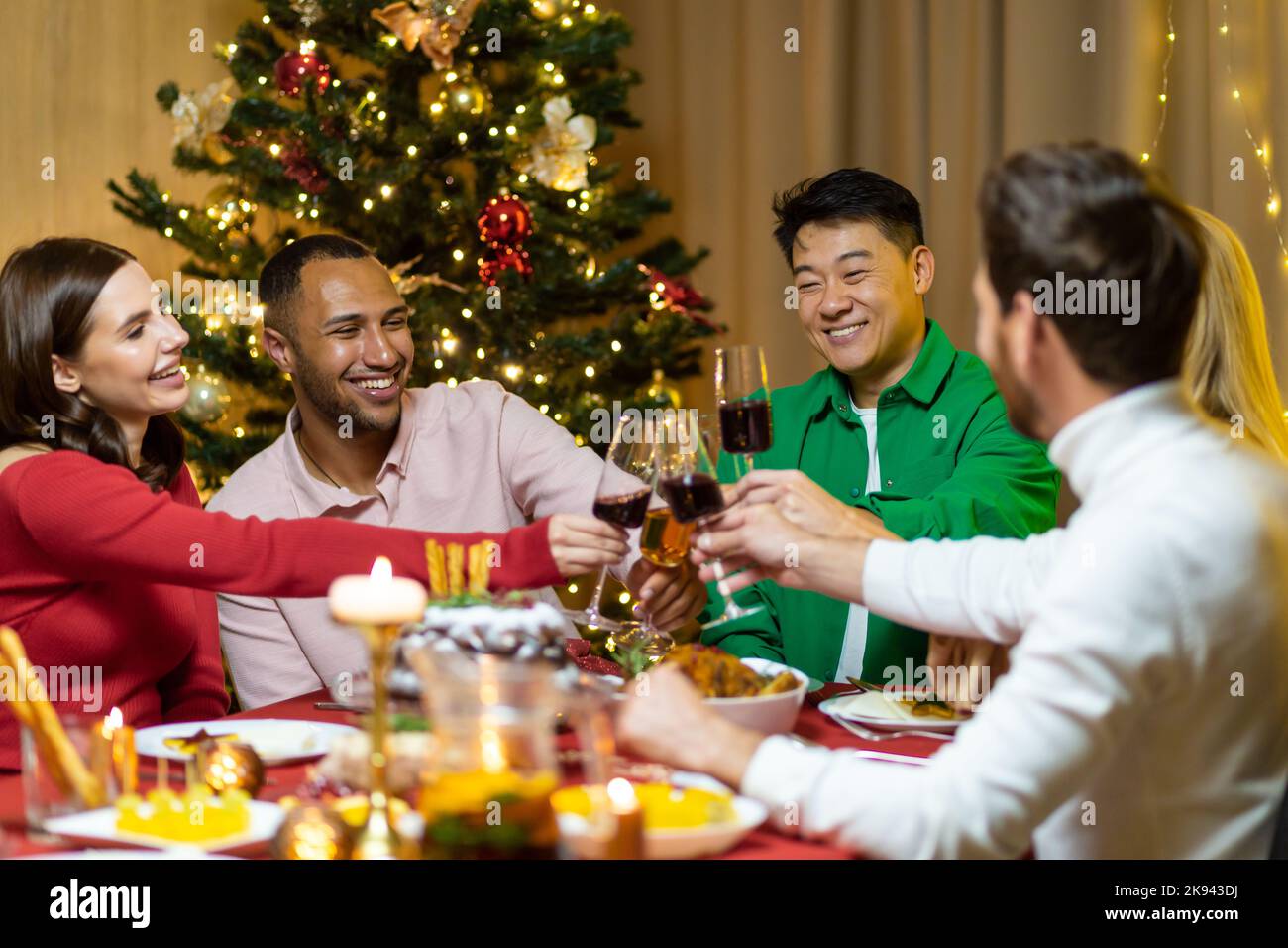 Festive Christmas dinner, diverse friends celebrating new year at home, eating and drinking wine, smiling and wishing each other happy holidays. Stock Photo
