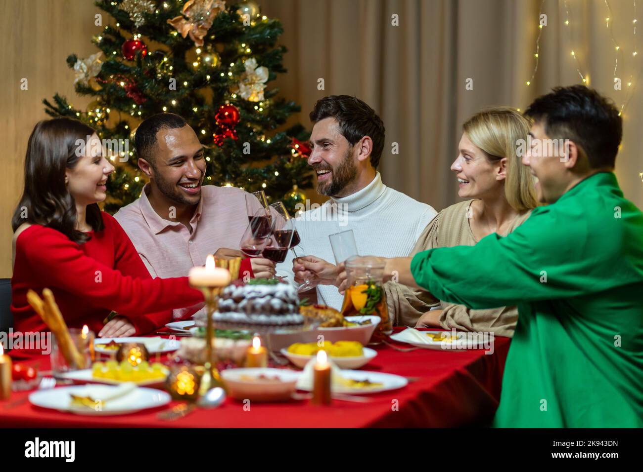 Festive Christmas dinner, diverse friends celebrating new year at home, eating and drinking wine, smiling and wishing each other happy holidays. Stock Photo