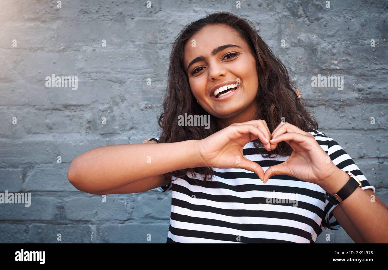 My heart is packed full of love for you. Portrait of a young woman making a heart shape with her hands against a grey wall. Stock Photo