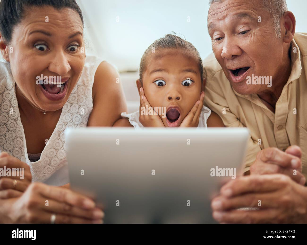 Tablet, family and shocked face child look at video online bonding together at home. Wow grandparents, young girl and fun activity streaming movie or Stock Photo