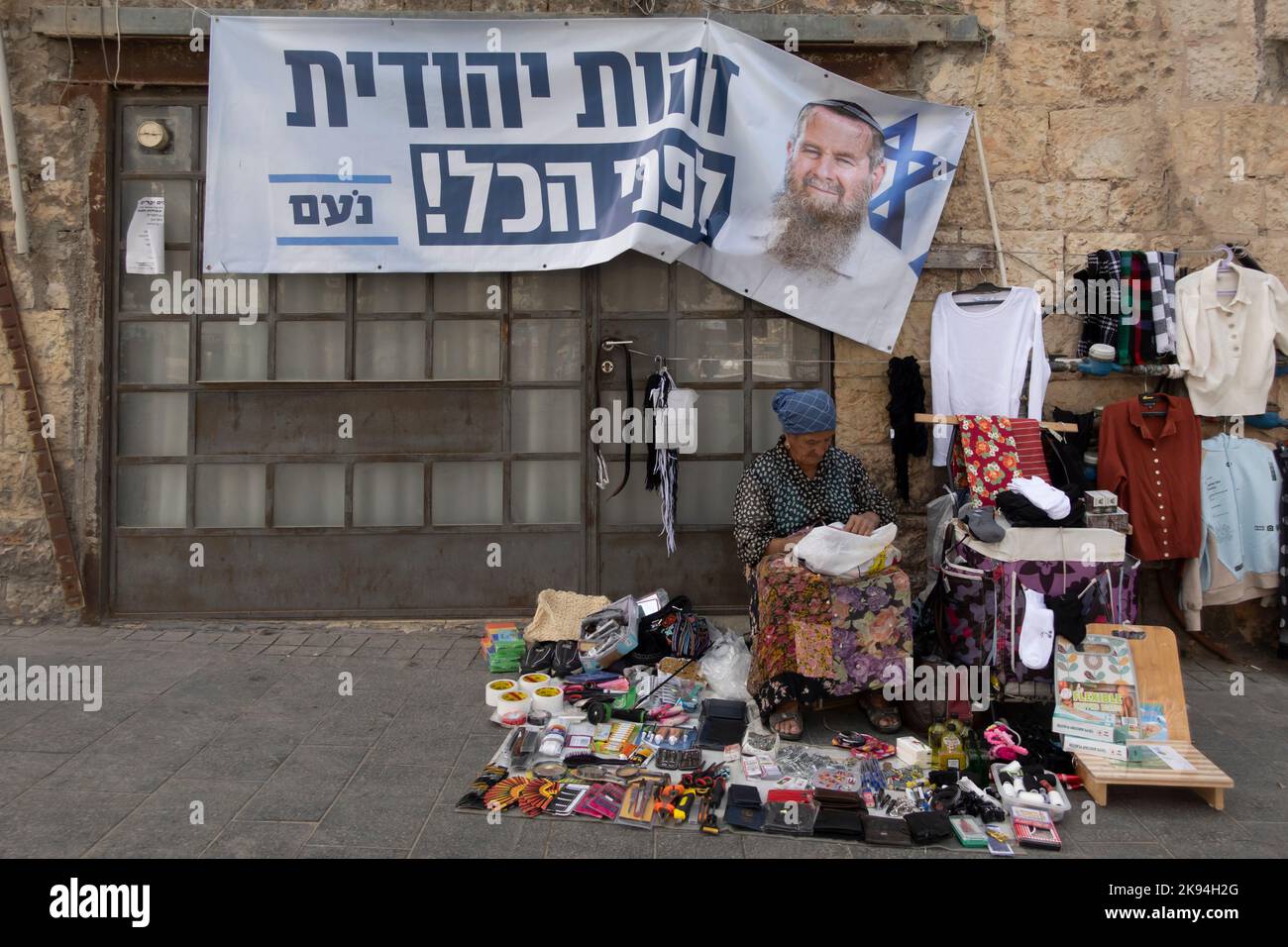 An Israeli street vendor sits beneath an election campaign poster for the  far-right Orthodox Jewish political party Noam depicting its leader Avi Maoz with a a caption reading  'Jewish identity above all'  is displayed in Jaffa road on October 25, 2022 in Jerusalem, Israel. Public opinion polls show significant support for the Religious Zionism party running together with the far-right Jewish Power ahead of parliamentary elections set for November 1 Stock Photo