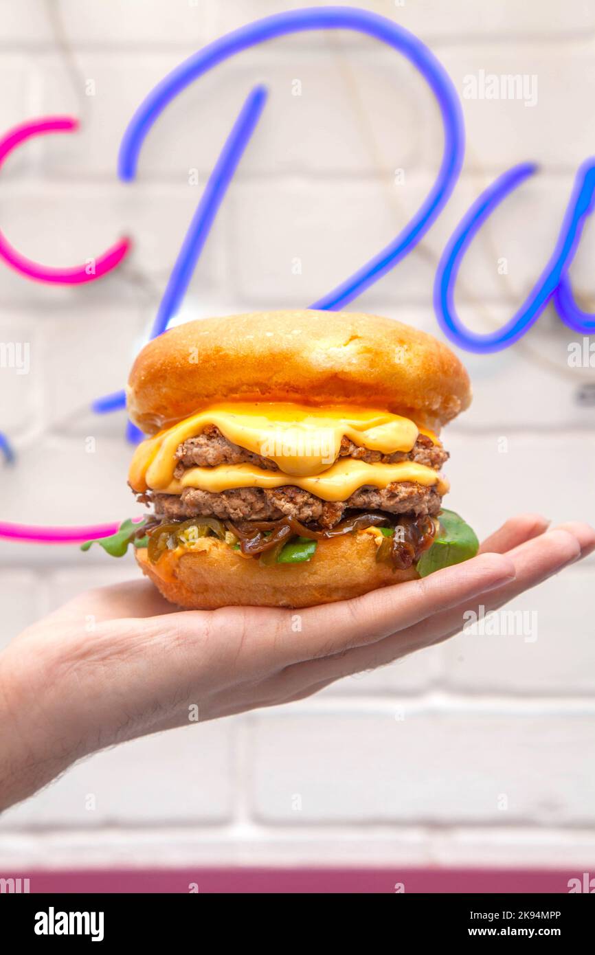 Hand palm holding a quesso melted cheese burger against a wall in cafe with neon sign. Stock Photo