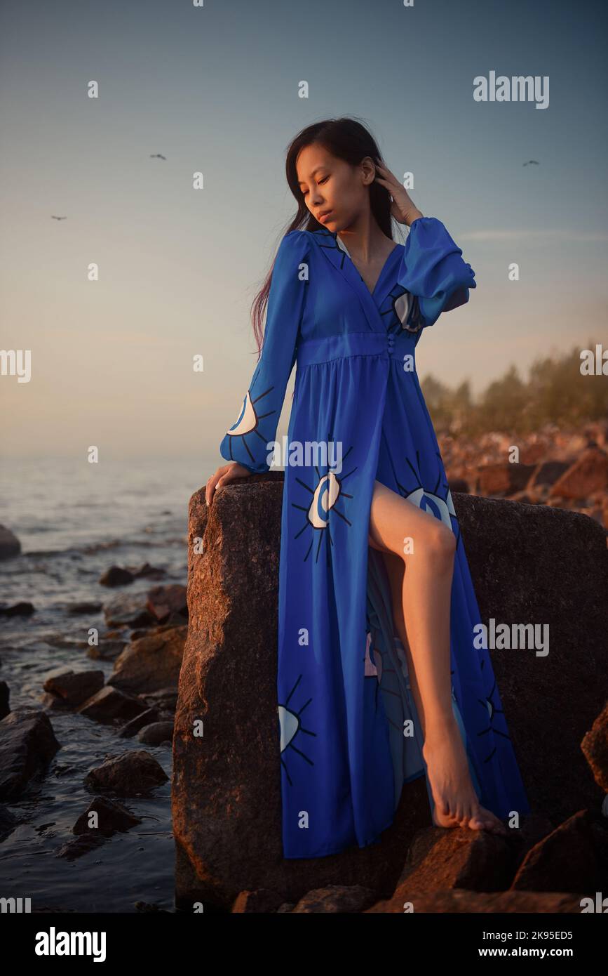 Young beautiful asian woman in long blue dress sitting on rock outdoors Stock Photo