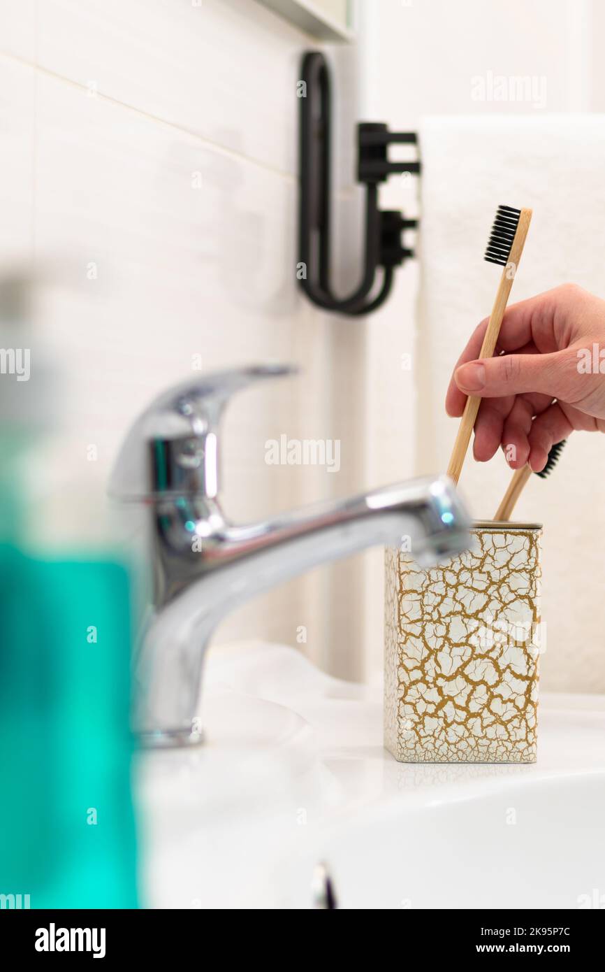A woman's hand puts a wooden toothbrush into a glass with a gold pattern in the bathroom against the backdrop of a faucet and glossy tiles. Selective Stock Photo