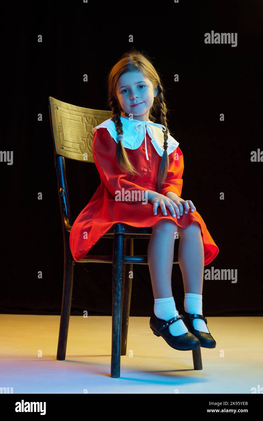 Portrait of beautiful cute little girl with pigtails wearing red festive dress sitting on chair isolated over dark background. Concept of children Stock Photo