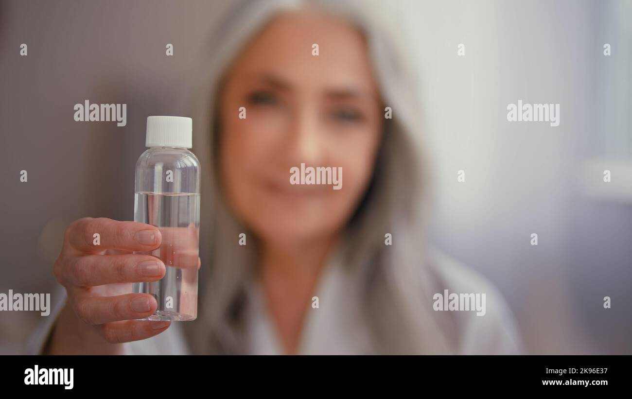 Old gray-haired elderly lady Caucasian woman in bathrobe shows advertises bottle with tonic serum oil with vitamins collagen minerals micellar water Stock Photo
