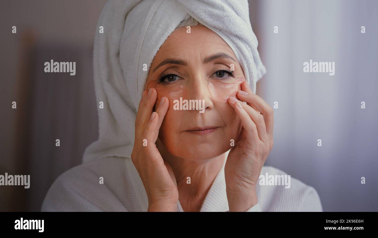 Close up portrait happy senior lady with anti wrinkle skin hydrogel collagen patches under eyes smiling to camera old 60s Caucasian woman with towel Stock Photo