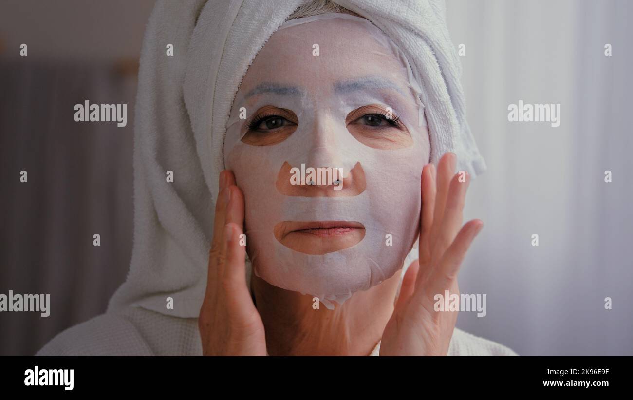 Portrait in bath old senior mature Caucasian woman wrap towel on head grandmother lady apply skincare textile skin mask on female face facial care Stock Photo