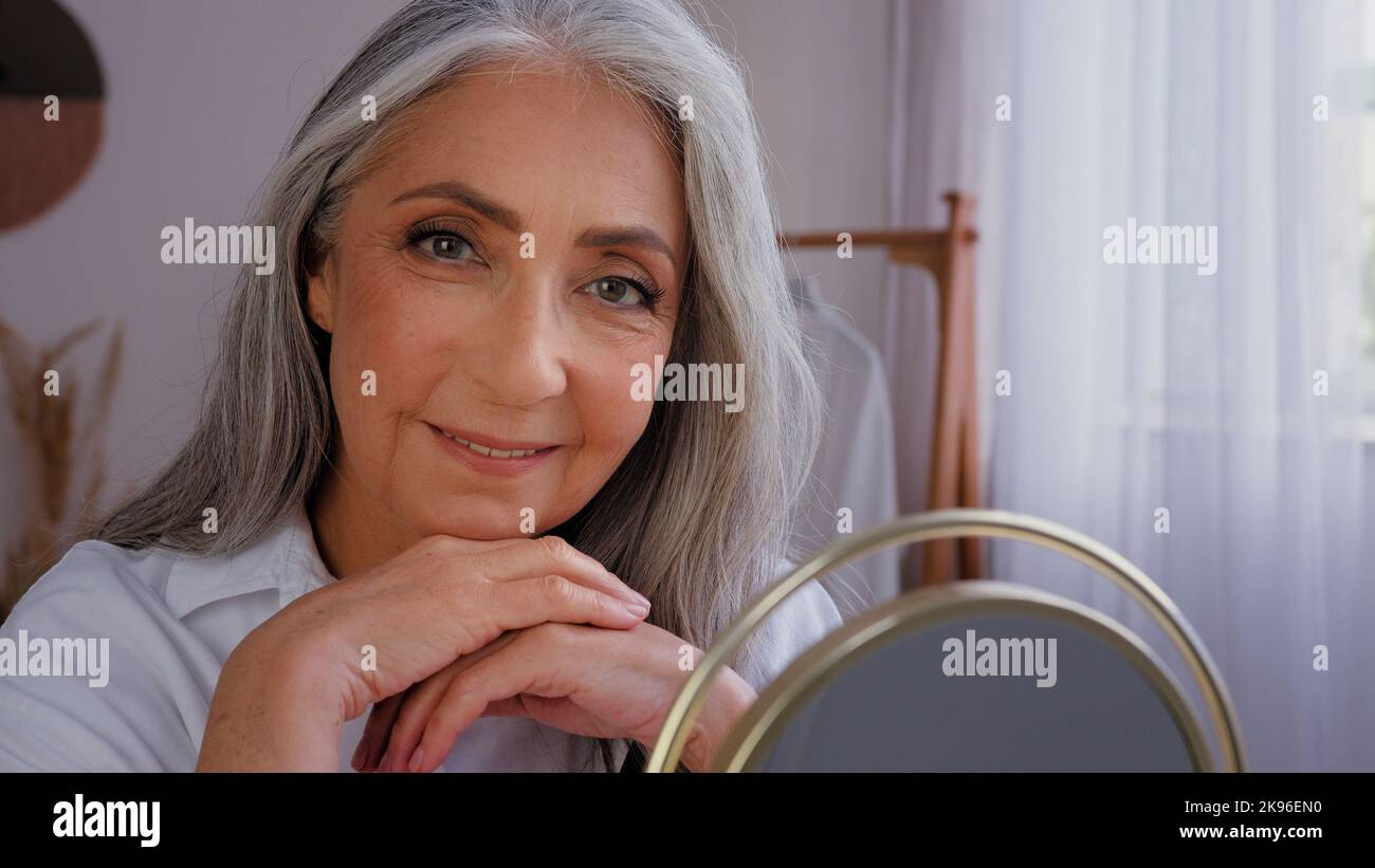 Senior gray-haired older 60s woman 50s lady granny with wrinkled beautiful aging face with perfect smooth skin looking at mirror reflection touching Stock Photo