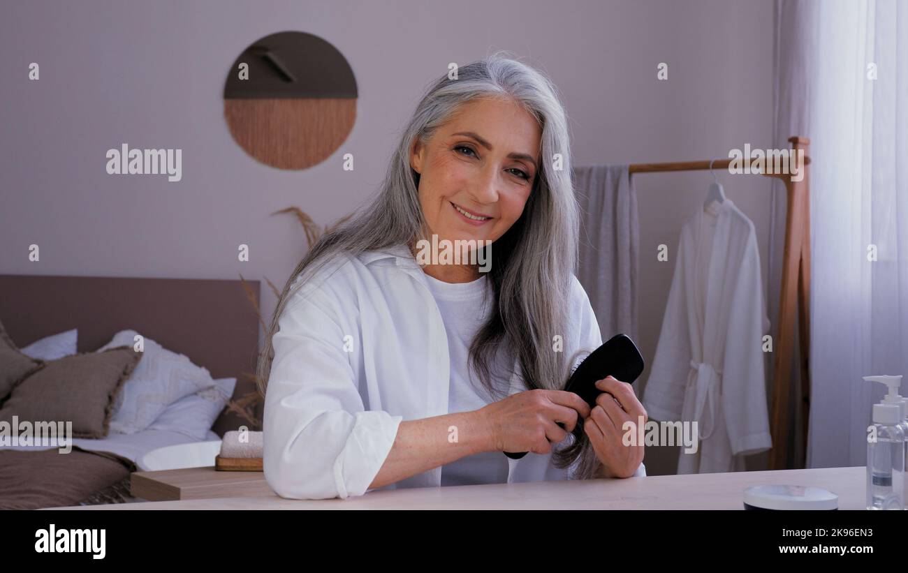 Portrait elderly mature middle-aged lady old 50s aged Caucasian woman in bathrobe morning beauty routine looking at camera combing gray long shiny Stock Photo