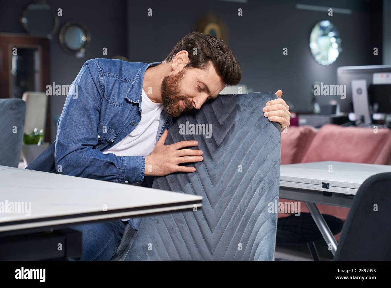Attractive bearded man leaning to soft chair back, while exploring linear pattern before buying furniture. Handsome guy checking texture of furniture textile, while touching chair. Concept of design. Stock Photo