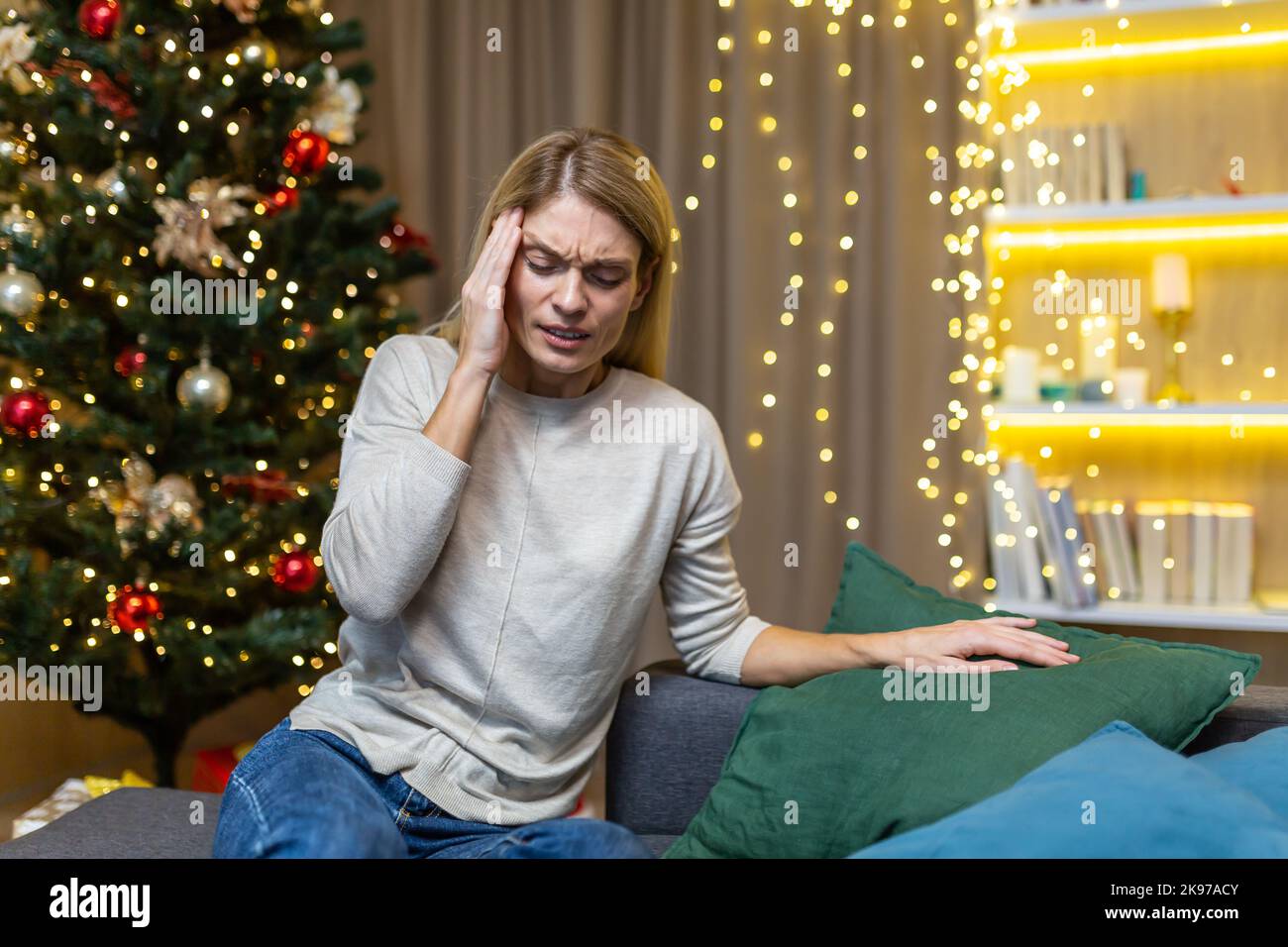 Sick woman on Christmas sitting on the sofa in the living room has a severe headache near the Christmas tree, migraine on the New Year holidays. Stock Photo