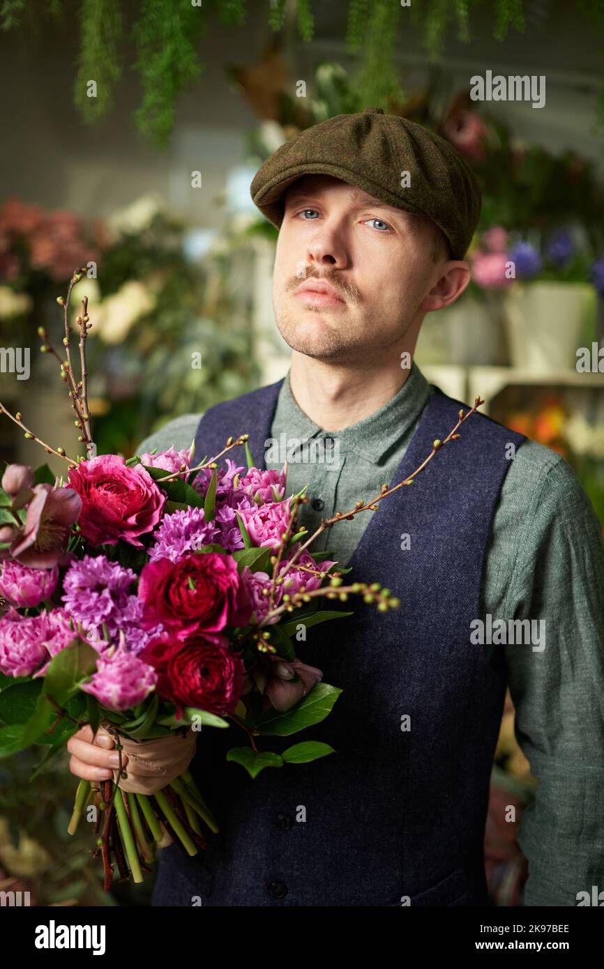 Mother's Day or Valentine's Day concept. Serious emotion: caucasian male in peaked cap and vintage 20s clothes holding bouquet of red and violet flowers. Congratulating with flowers Stock Photo