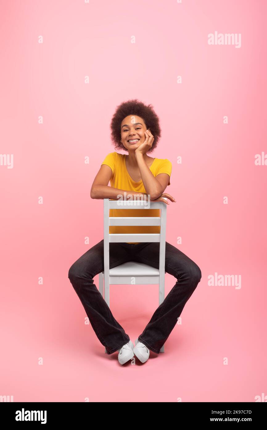 Smiling pretty young mixed race woman with curly hair in casual sits on chair, isolated on pink background Stock Photo