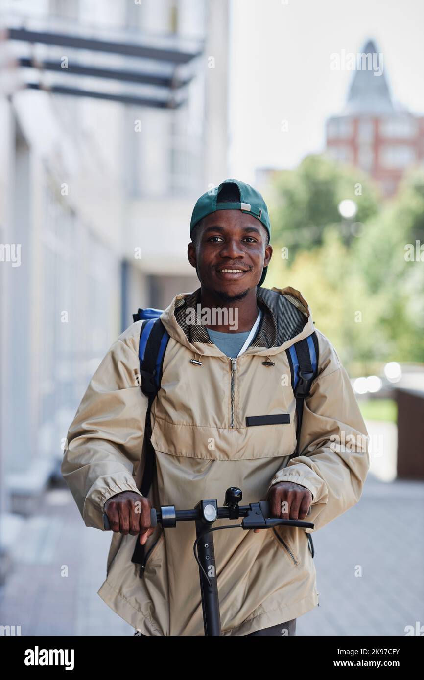 Vertical portrait of smiling food delivery boy riding electric scooter in city street Stock Photo