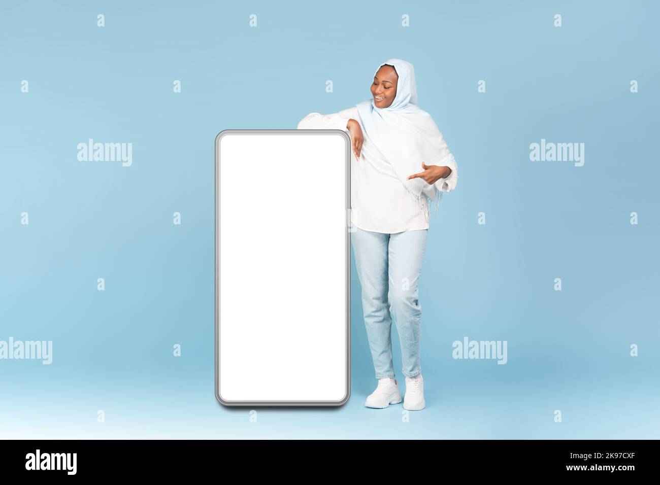 Wow, great offer. Full body length of happy black muslim woman pointing at big blank smartphone display, mockup Stock Photo