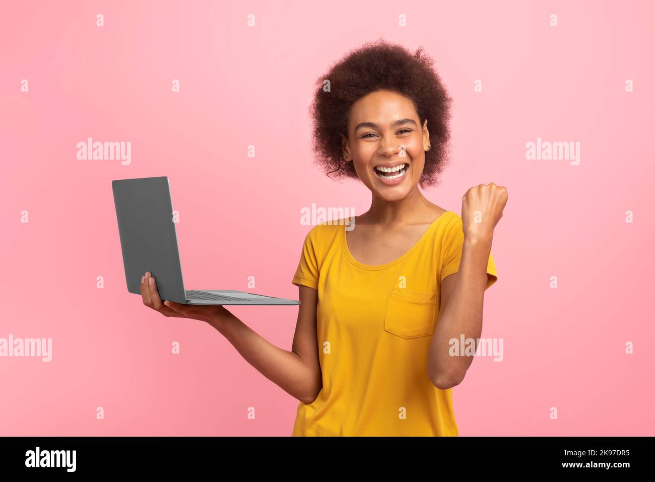 Cheerful excited young mixed race woman with curly hair rejoices online success with laptop, making victory gesture Stock Photo