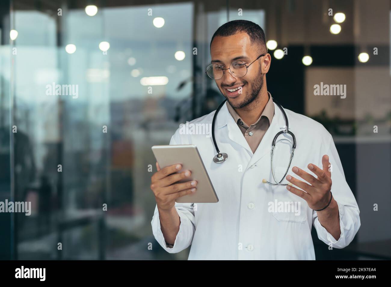 A young handsome male hispanic doctor is having an online consultation with a patient. Standing in the hospital with a stethoscope, holding a tablet in his hands, explaining with his hands. Stock Photo