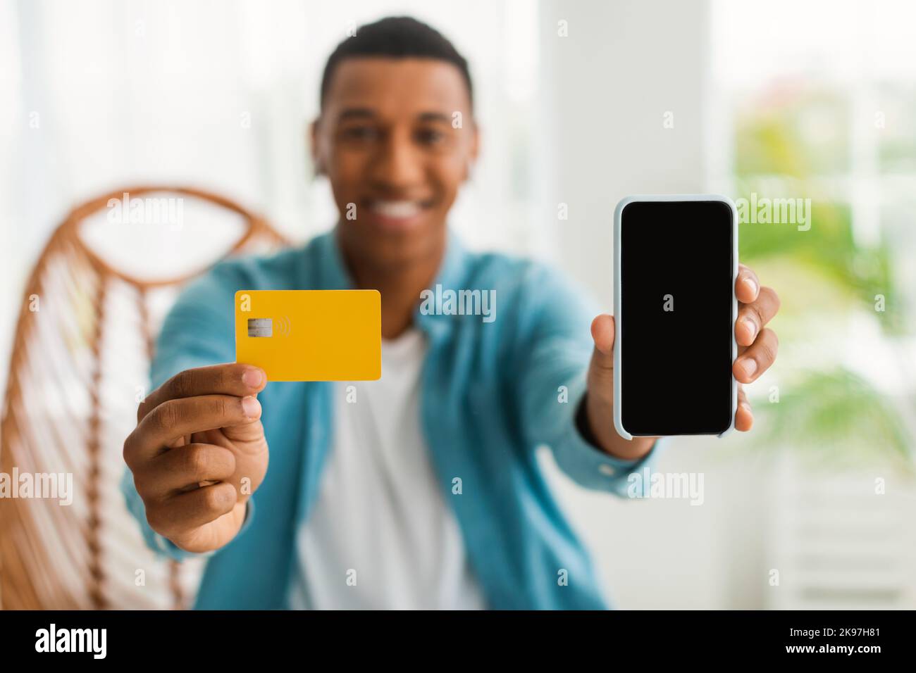 Cheerful young black male shopaholic in casual showing credit card and smartphone with blank screen Stock Photo