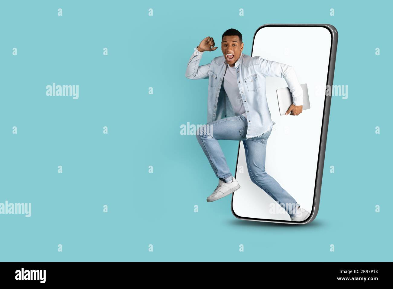 Cheerful excited millennial black man with open mouth with laptop jumping out of huge smartphone Stock Photo