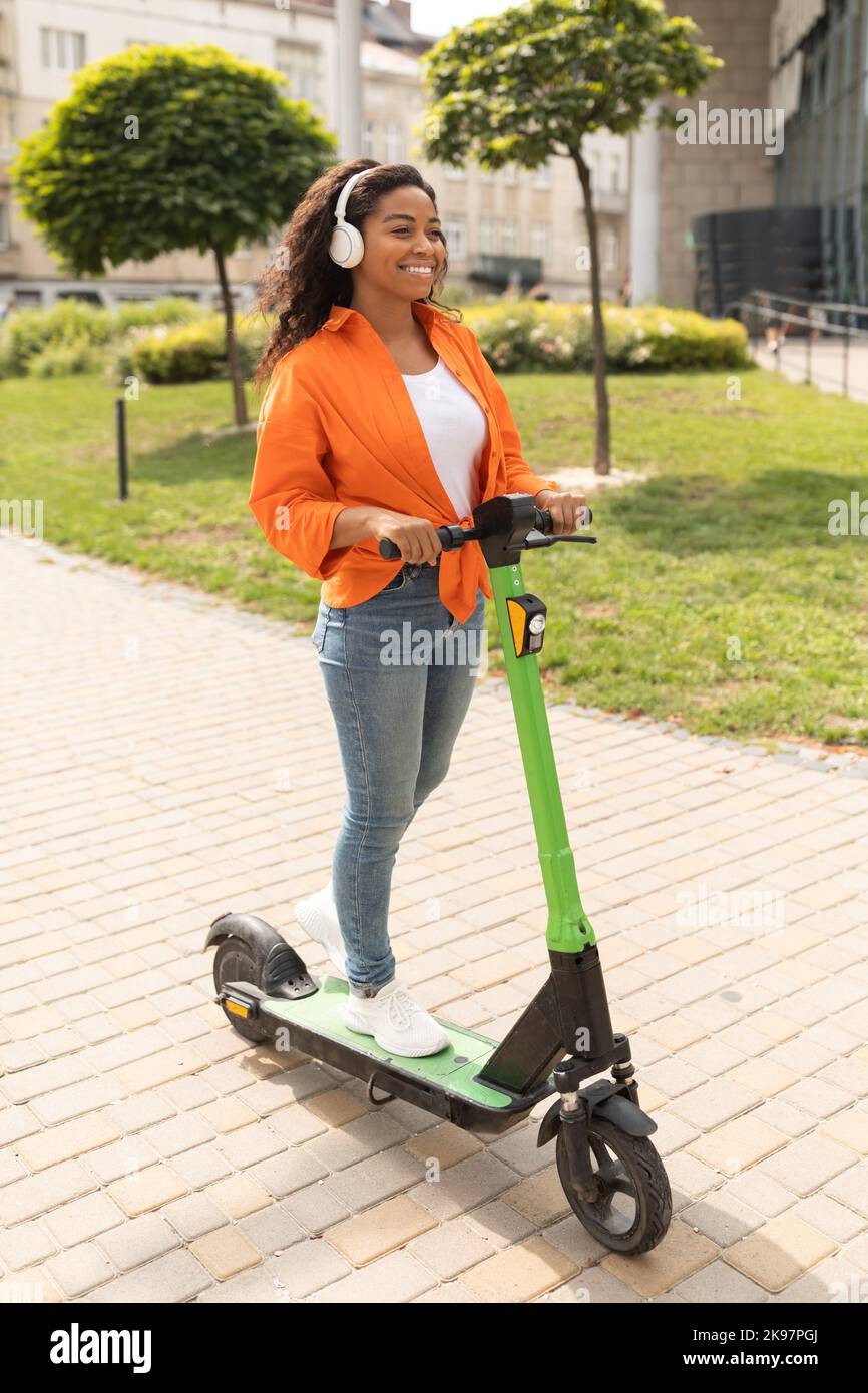 Happy millennial african american lady in casual, wireless headphones, rides on electric scooter Stock Photo