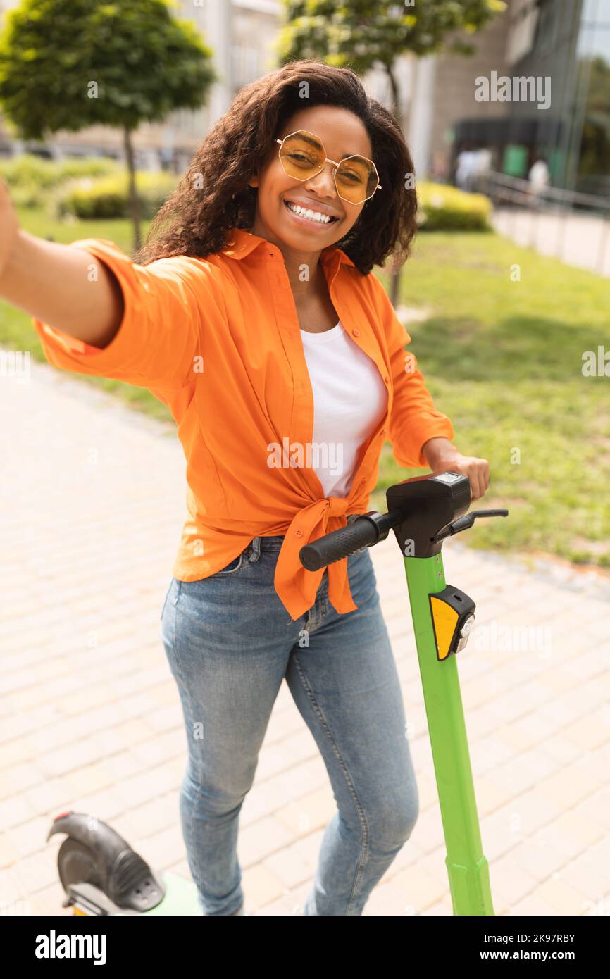 Happy millennial african american lady in casual taking selfie on smartphone on electric scooter in city street Stock Photo
