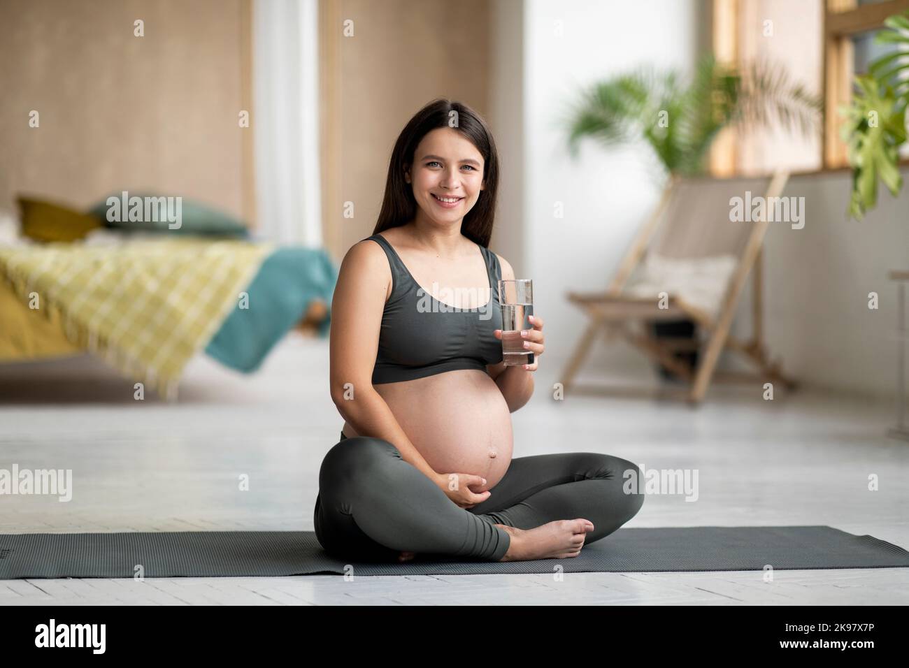 Young Pregnant Woman Sitting On Yoga Mat And Holding Glass With Water Stock Photo
