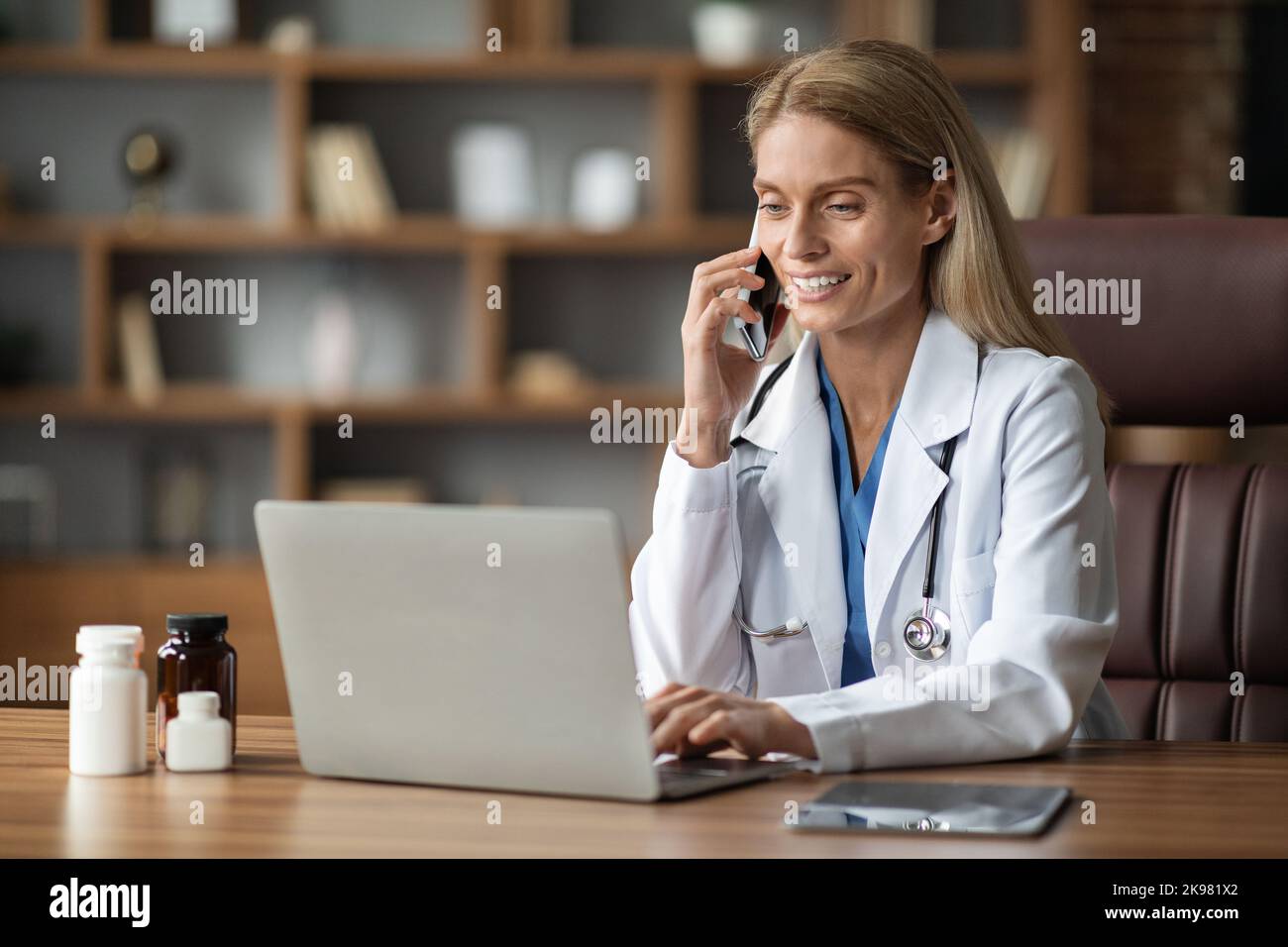 Smiling Doctor Woman Talking On Cellphone And Using Laptop At Workplace Stock Photo