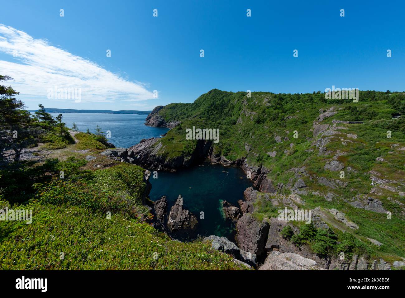 A rugged ocean coastline with high rocky sea stacks, cliffs, vibrant green grass, blue sky, and clouds. The smooth water is a blue color. Stock Photo