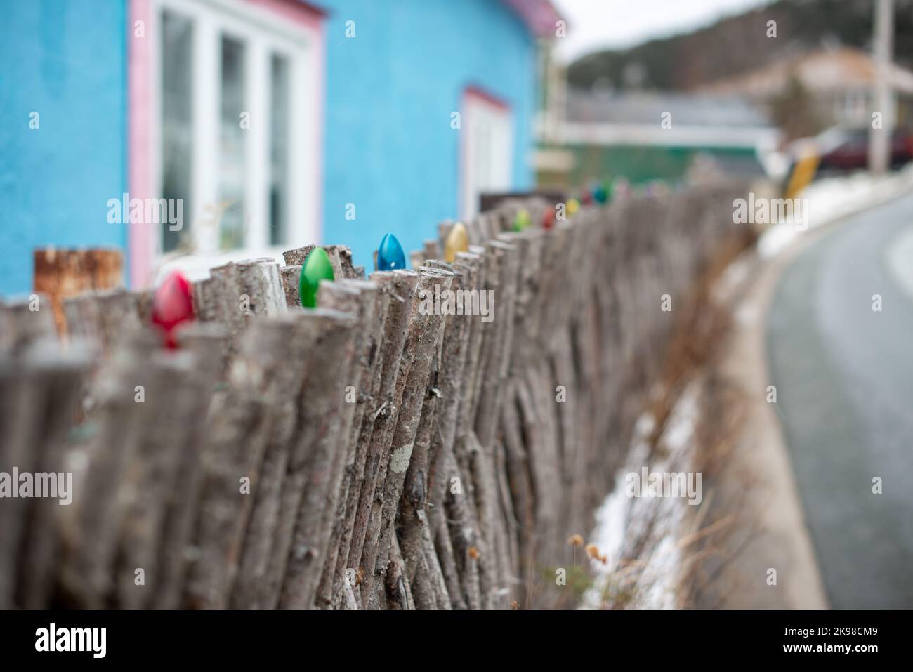 A rustic wooden interlocking twig fence with multicolored Christmas lights on the top. A road on one side and a blue house behind. Stock Photo