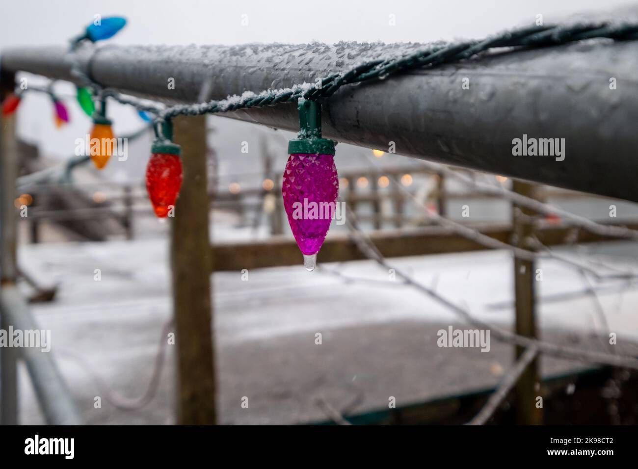 A string of colorful Christmas lights strung around a black metal handrail. There's fresh white snow on the rail and concrete ground. Stock Photo