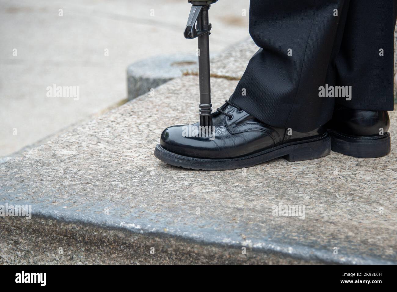 A  military person stands at attention with his rifle pitched on the toe of his shinned boot. The male navy officer stands on guard wearing dark pants Stock Photo