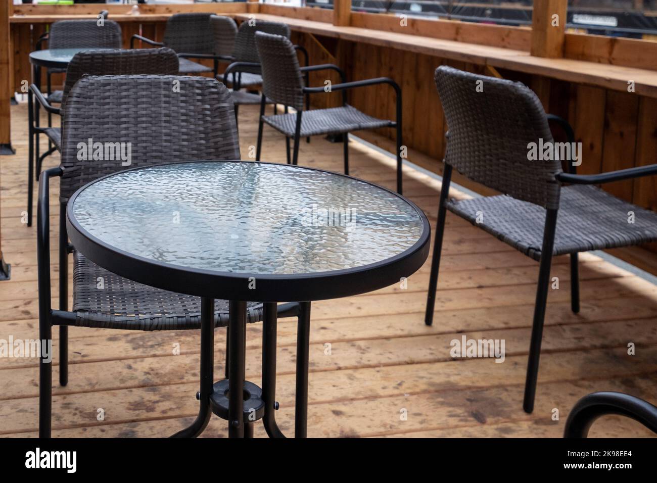 Multiple empty black metal tables with round glass tops and black rattan chairs on a sidewalk cafe patio of an outdoor restaurant. Stock Photo