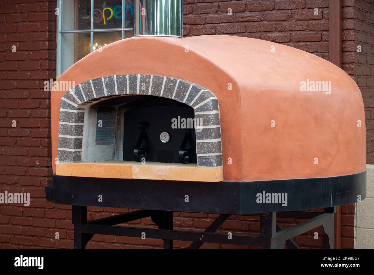 An exterior of a gas clay pizza oven on black metal legs on a restaurant patio. There are bricks surrounding the opening of the Italian equipment. Stock Photo